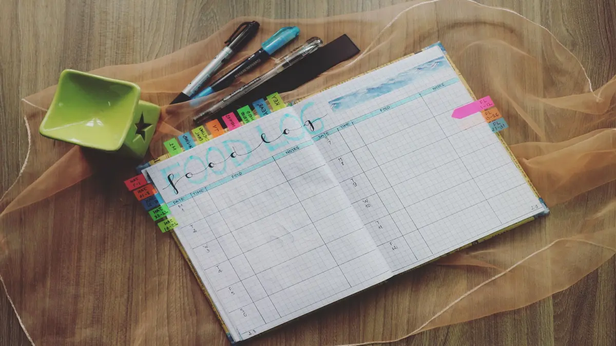 Open planner with color-coded tabs, pens, and a green cup on a wooden desk.