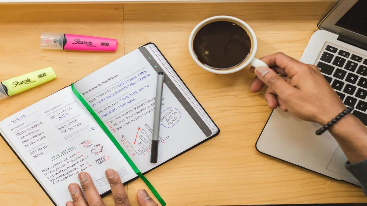 Person taking notes in a notebook with coffee and a laptop on the desk.
